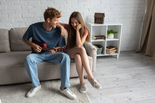 Full length of curly and redhead young man playing ukulele near happy girlfriend in living room - foto de stock