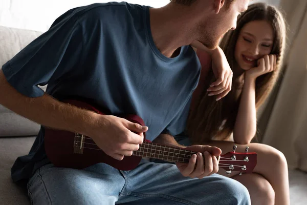 Молодий чоловік грає укулеле біля щасливої дівчини у вітальні — стокове фото