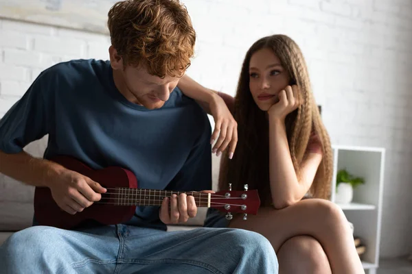 Curly and redhead young man playing ukulele near blurred girlfriend in living room - foto de stock