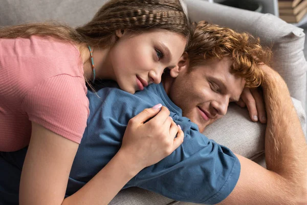 High angle view of pretty teenage girl lying on back of smiling boyfriend and resting on couch — Stock Photo