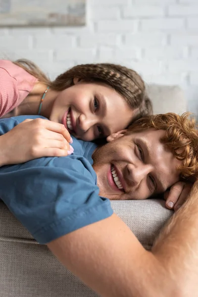 Joyful teenage girl lying on back of smiling boyfriend and resting on couch — Stock Photo