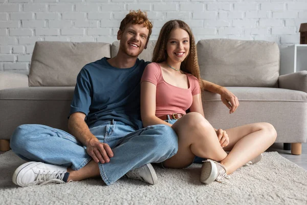 Happy young couple smiling while sitting near couch in modern living room — стоковое фото