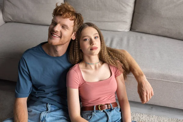 High angle view of cheerful young man sitting with girl near couch in modern living room - foto de stock