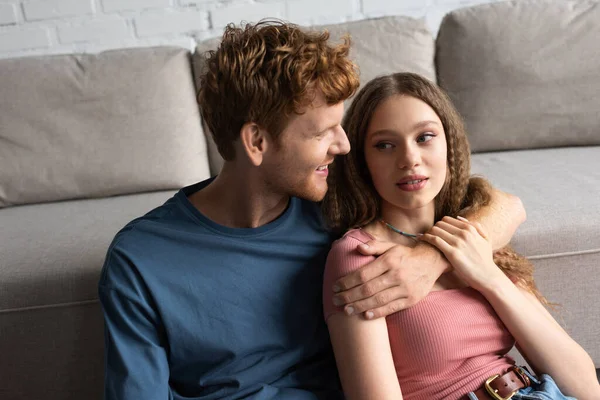 Redhead and happy young man hugging and looking at pretty girlfriend in living room - foto de stock