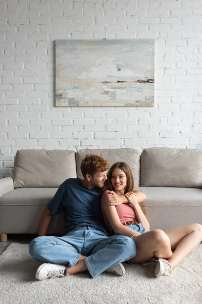 Redhead and happy young man hugging and looking at cheerful girlfriend in modern living room — Photo de stock