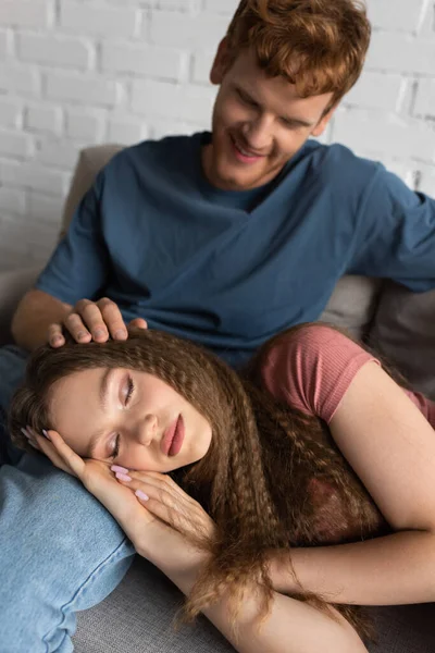 Redhead and happy young man stroking hair of sleepy girlfriend in living room - foto de stock