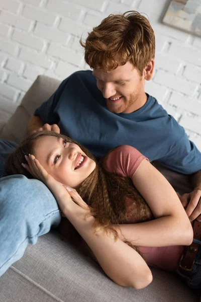 Redhead and smiling young man stroking hair of cheerful girlfriend in living room — стоковое фото