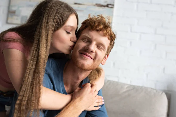 Pretty teen girl hugging and kissing cheek of redhead boyfriend in living room — Stockfoto