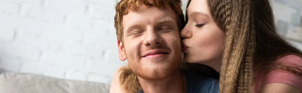 Pretty teen girl hugging and kissing cheek of redhead boyfriend in living room, banner — стоковое фото