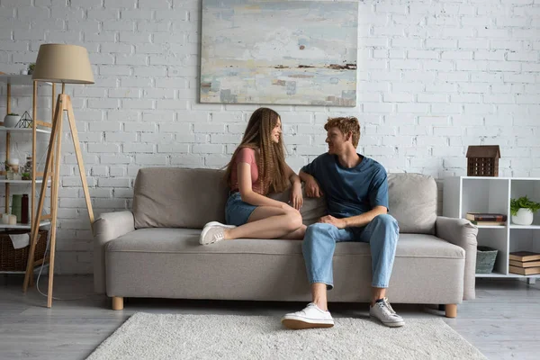 Full length of young and happy couple sitting on couch and looking at each other in living room — стоковое фото