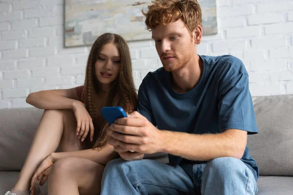 Young redhead man using smartphone while sitting near girlfriend on couch — стоковое фото