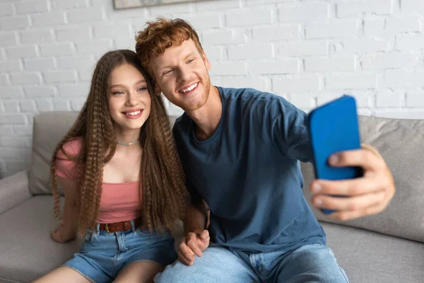 Young redhead man taking selfie with happy girlfriend in living room - foto de stock
