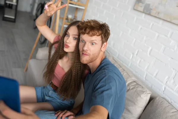 Young redhead man taking selfie with girlfriend pouting lips and showing peace sign in living room — Fotografia de Stock