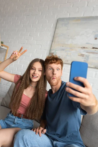 Young redhead man grimacing and taking selfie with happy girlfriend showing peace sign in living room — Stockfoto