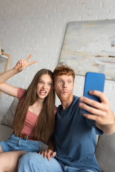 Young redhead man grimacing and taking selfie with girlfriend showing peace sign in living room — Stockfoto