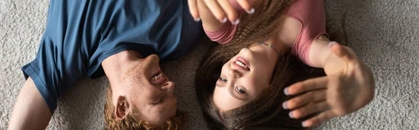 Top view of happy and young man lying on carpet with cheerful girlfriend smiling while gesturing in living room, banner — Photo de stock