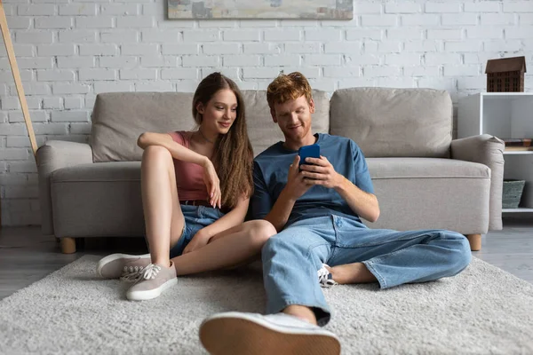 Young redhead man using smartphone while sitting near smiling girlfriend and couch in living room — стоковое фото