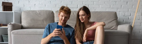 Young redhead man using smartphone while sitting near smiling girlfriend and couch in living room, banner - foto de stock