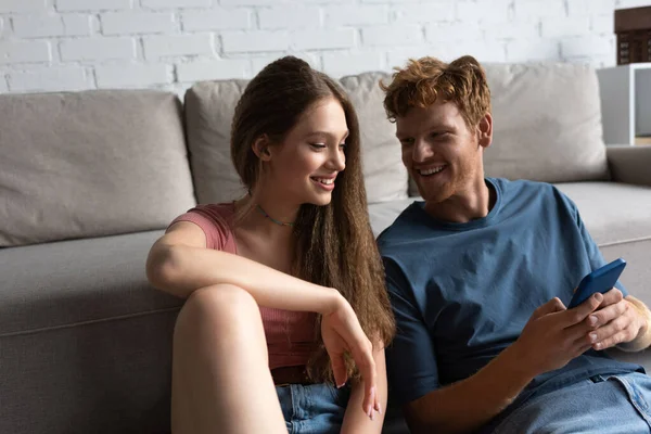 Young redhead man holding smartphone while looking at smiling girlfriend in living room — Foto stock