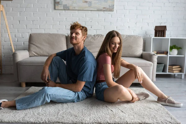 Full length of happy young couple sitting on carpet in modern living room — Photo de stock