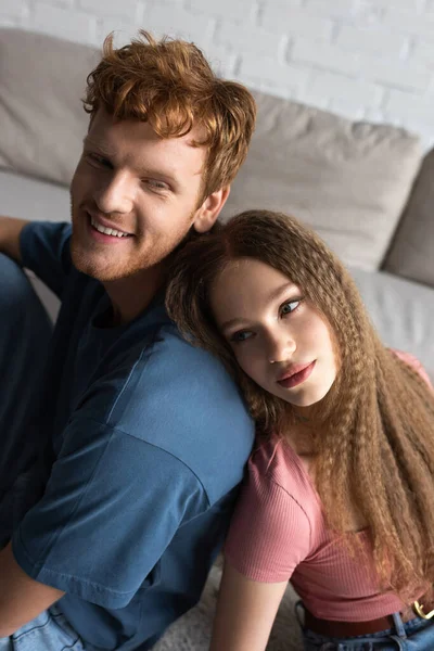 Teenage girl leaning on back of happy boyfriend with red hair in living room - foto de stock