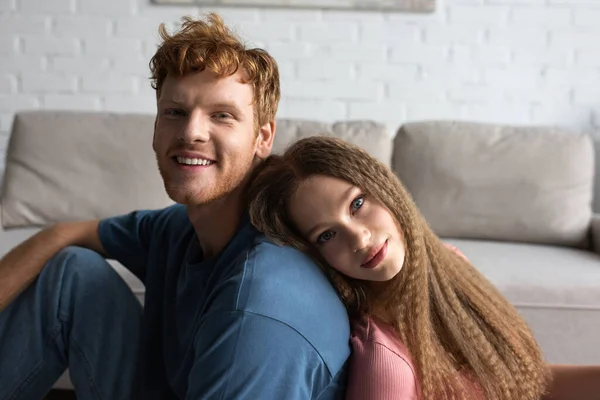 Teenage girl leaning on back of cheerful boyfriend with red hair in living room - foto de stock