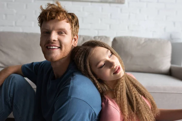 Pretty teenage girl leaning on back of positive boyfriend with red hair in living room — Fotografia de Stock