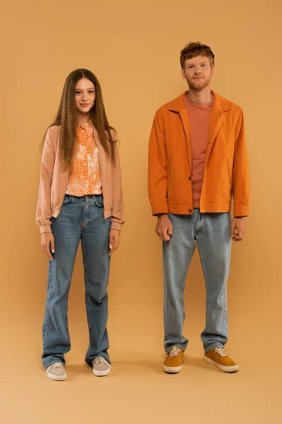 Full length of happy young couple in autumnal outfits and gumshoes standing together on beige — стоковое фото