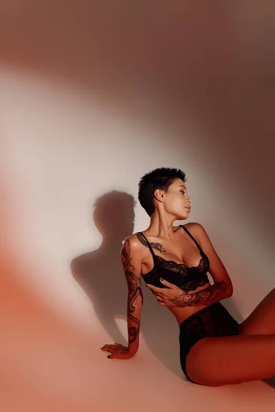 Татуированная брюнетка женщина в сексуальном белье сидит на красном фоне с тенью — стоковое фото