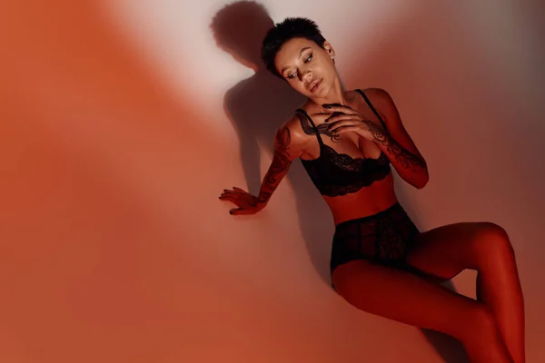 Visão de alto ângulo da mulher tatuada sensual em lingerie de renda preta no fundo vermelho e marrom — Fotografia de Stock