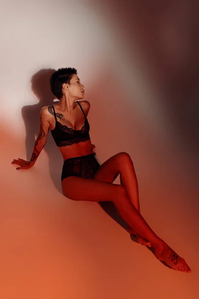 Longitud completa de mujer tatuada en ropa interior sexy sentado sobre fondo rojo pálido con sombra - foto de stock