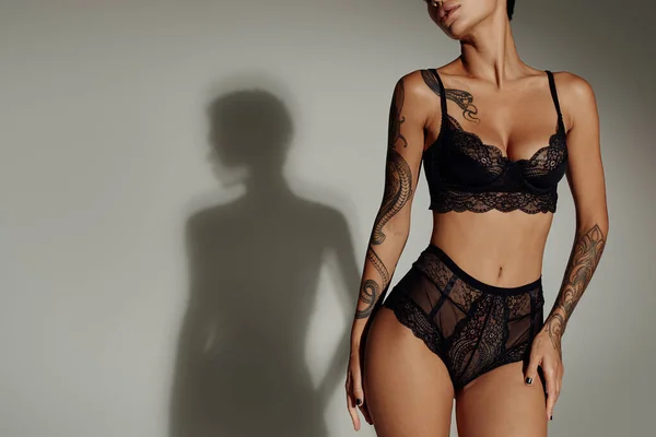 Cropped view of tattooed woman in black lace lingerie on grey background with shadow — Stock Photo