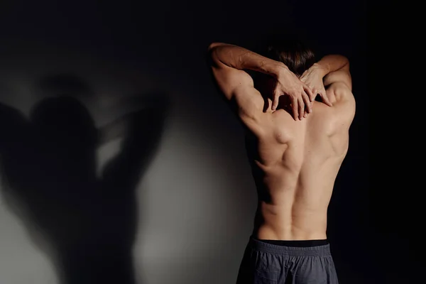 Vista posterior del hombre musculoso sin camisa de pie sobre fondo negro y gris con sombra - foto de stock
