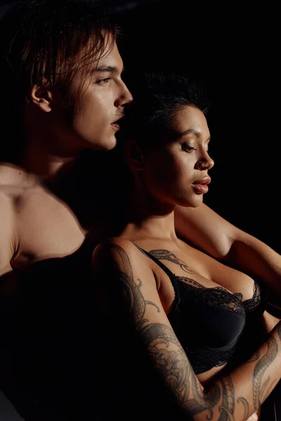 Side view of tattooed woman with sexy breast near shirtless man isolated on black — Stock Photo