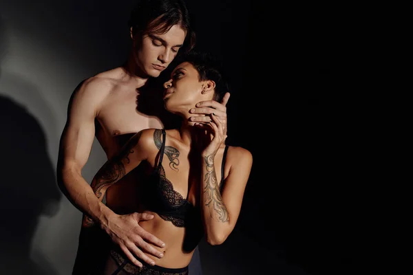 Молодой мужчина без рубашки, обнимающий страстную татуированную женщину в лифчике на черном и сером фоне — стоковое фото