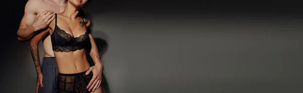 Abgeschnittene Ansicht eines Mannes, der tätowierte Frau in schwarzer Spitzenunterwäsche auf grauem Hintergrund umarmt, Banner — Stockfoto
