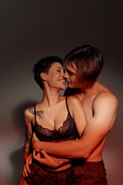 Mulher alegre e sexy no sutiã beijando com homem sem camisa abraçando-a no fundo cinza — Fotografia de Stock