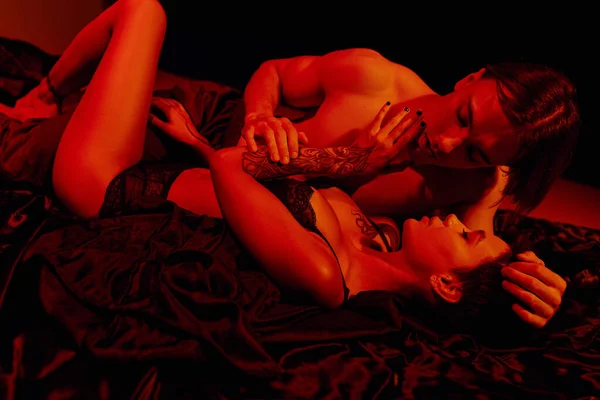 Sexy tattooed woman and young shirtless man embracing on black silk bedding in red light — Stock Photo