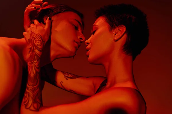 Вид збоку пристрасна татуйована жінка, що обіймає і цілує молодого чоловіка ізольовано на червоному — стокове фото