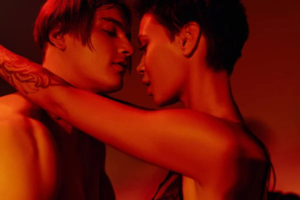 Вид сбоку сексуальной татуированной женщины, стоящей лицом к лицу с молодым человеком на красном фоне — стоковое фото