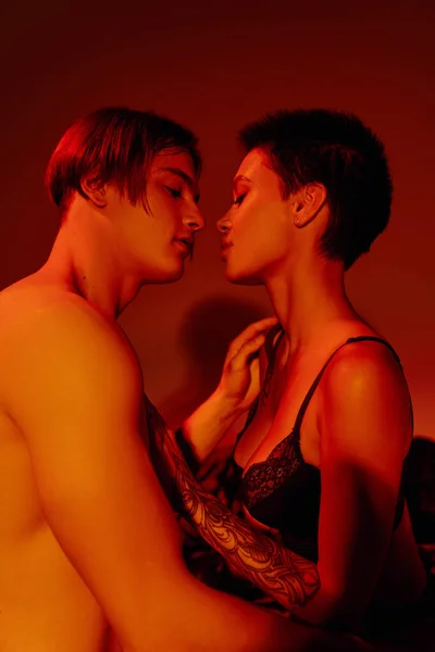 Вид сбоку сексапильного мужчины и соблазнительной женщины в черном бюстгальтере на красном фоне — стоковое фото