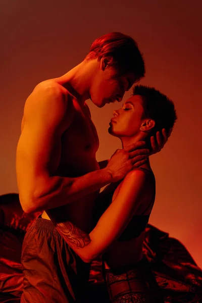 Vista lateral de un hombre joven sin camisa abrazando a una mujer apasionada en sujetador sobre fondo rojo - foto de stock