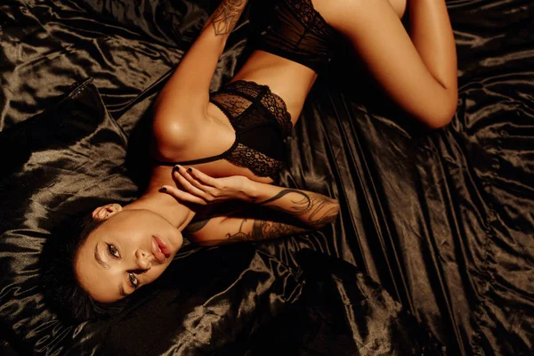 Top view of sexy tattooed woman in lace lingerie lying on black silk bedding and looking at camera — Stock Photo