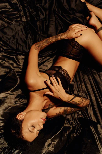 Top view of tattooed woman with sexy body lying in lace underwear on black satin bedding — Stock Photo