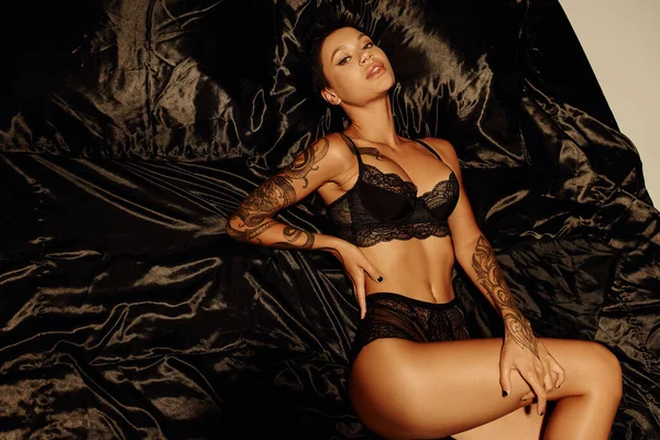 Vue de dessus de femme tatouée sexy en sous-vêtements noirs couché sur la literie en satin et regardant la caméra — Photo de stock