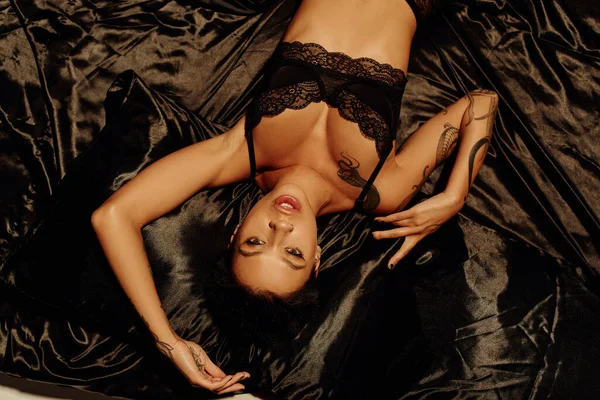 Vista superior de mulher tatuada apaixonado em sutiã de renda deitado em cama de cetim preto e olhando para a câmera — Fotografia de Stock