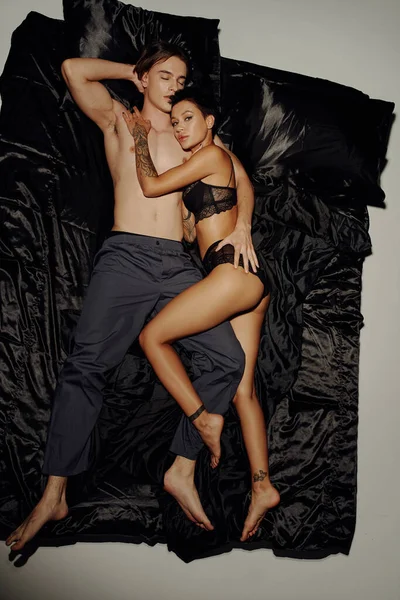 Vista superior da mulher tatuada em lingerie deitada na cama de seda preta perto do jovem e olhando para a câmera no fundo cinza — Fotografia de Stock