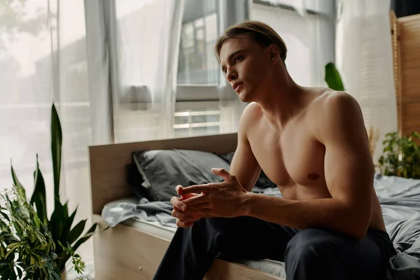 Giovane uomo senza maglietta in pigiama pantaloni seduto in camera da letto con piante decorative — Foto stock