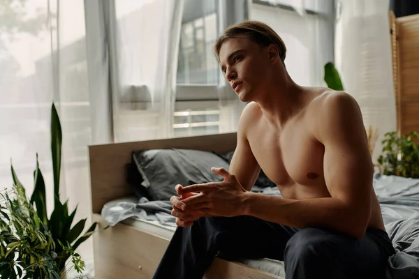 Young shirtless man in pajama pants sitting in bedroom with decorative plants — Stock Photo