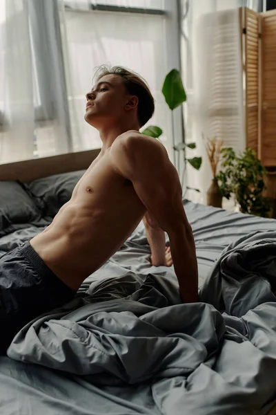 Vista lateral do homem jovem e sexy sentado na cama cinza em casa — Fotografia de Stock